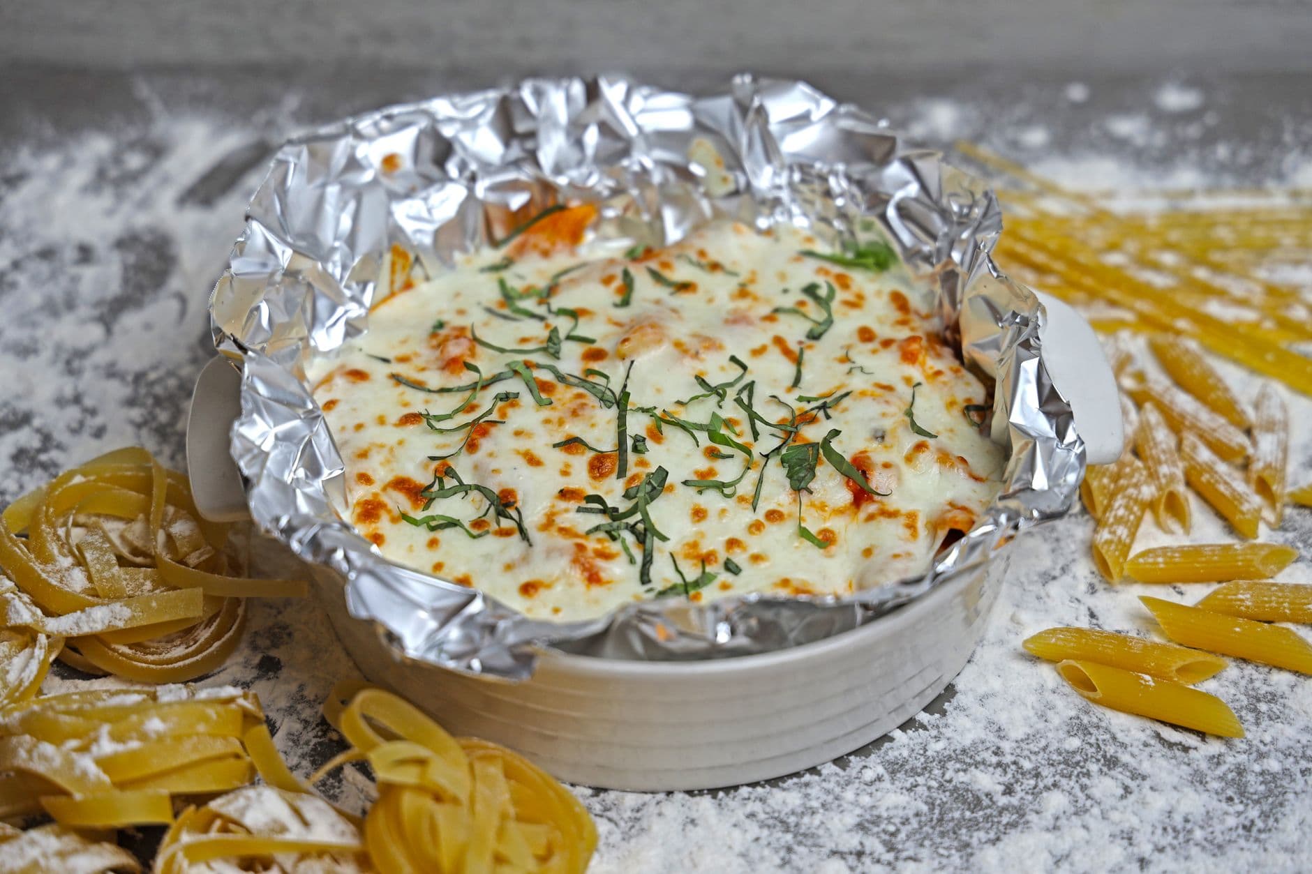 A mouthwatering image of baked pasta topped with cheese and fresh herbs surrounded by raw pasta and flour.
